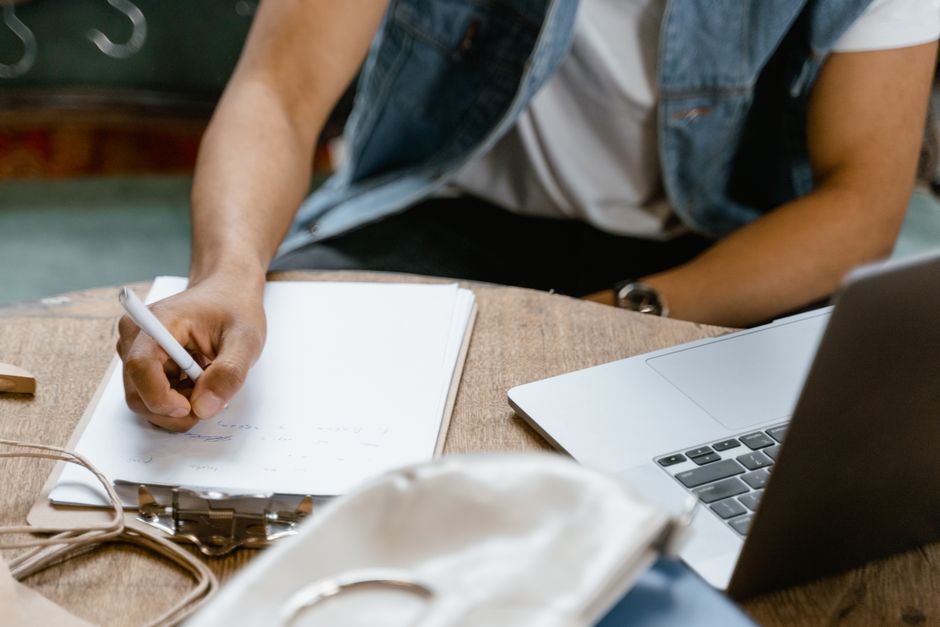 A person in denim vest writing notes on clipboard by laptop, indoors.
